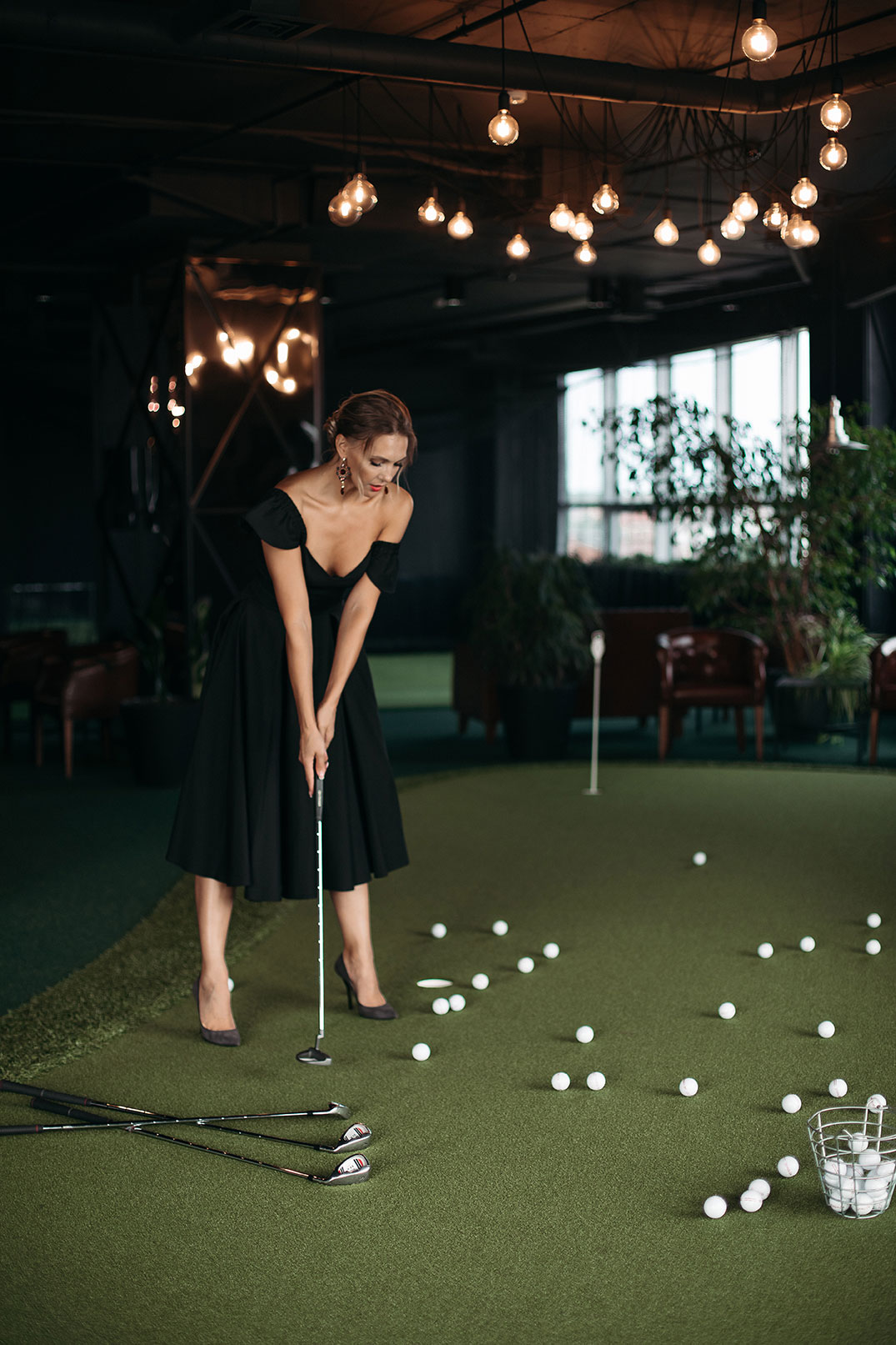 A woman in an evening gown and high heels stands on a synthetic turf mat, golf club in hand, and is surrounded by balls.