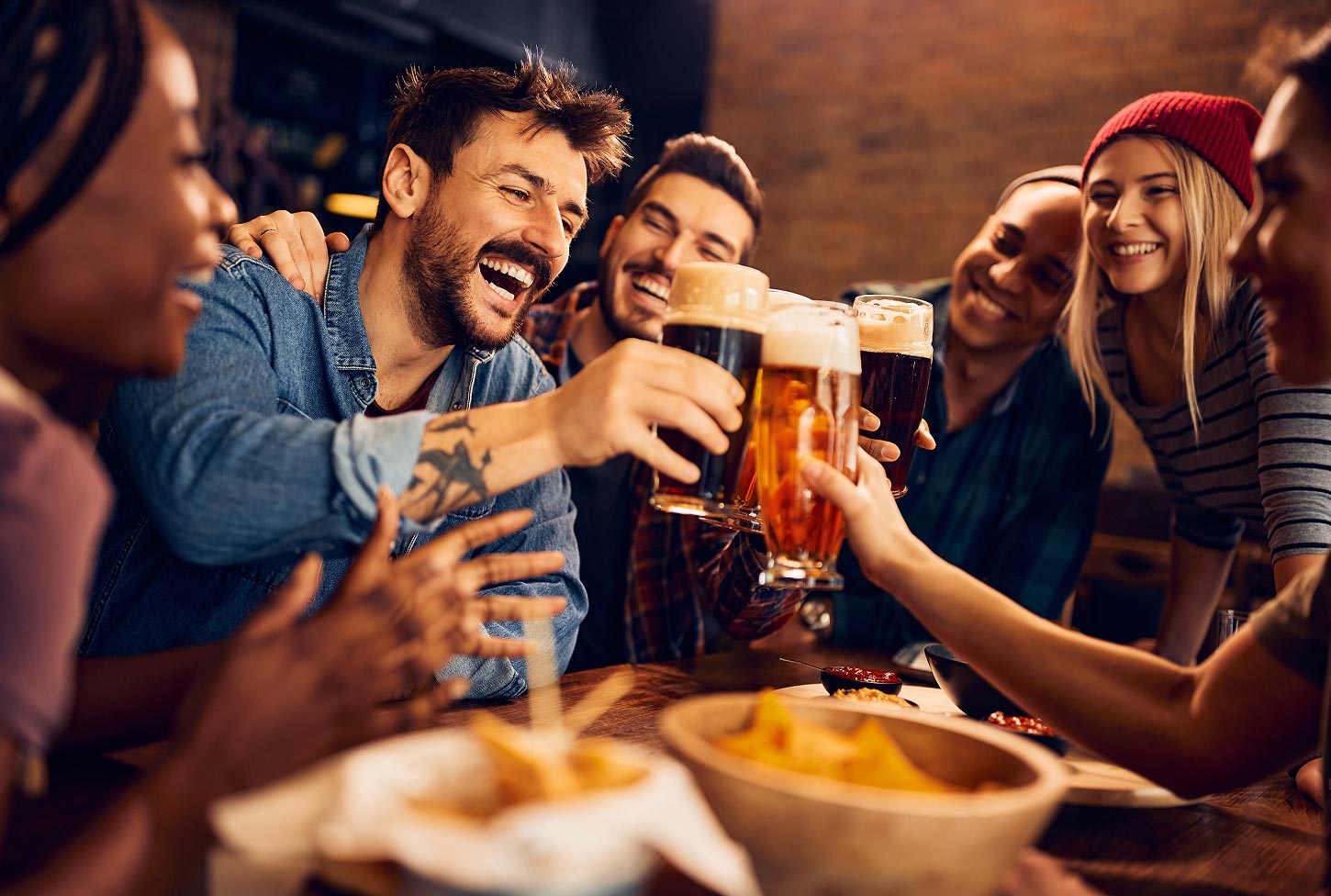 A group of friends smile and raise their glasses for a toast in a restaurant and bar, glasses of beer in hand.